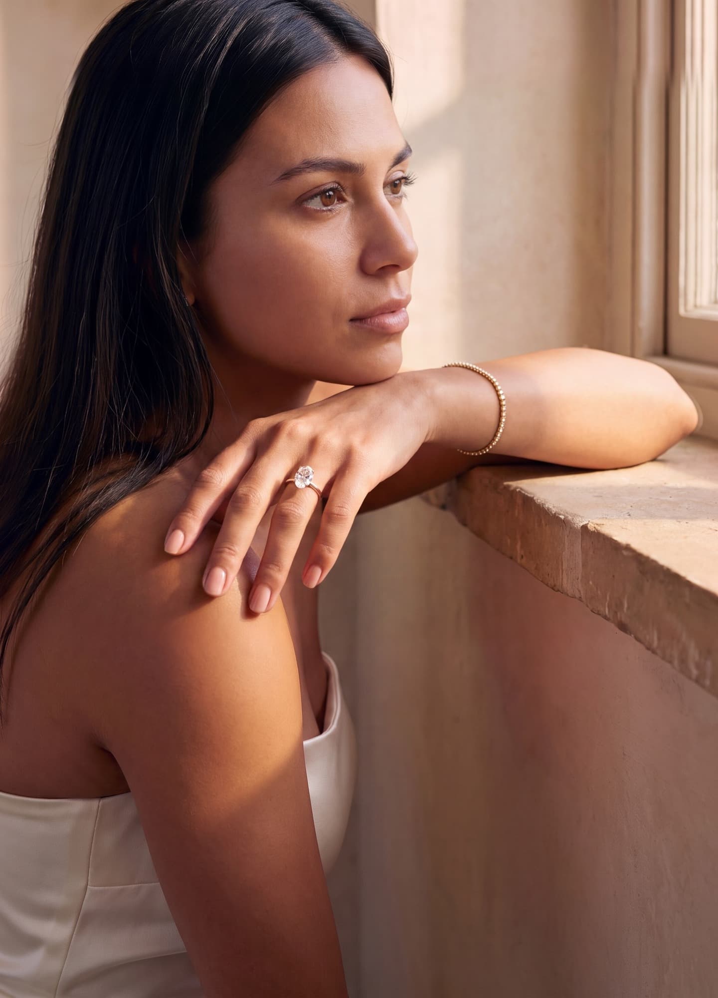 A woman by a sunlit window wearing an oval lab-grown diamond engagement ring and a diamond line bracelet