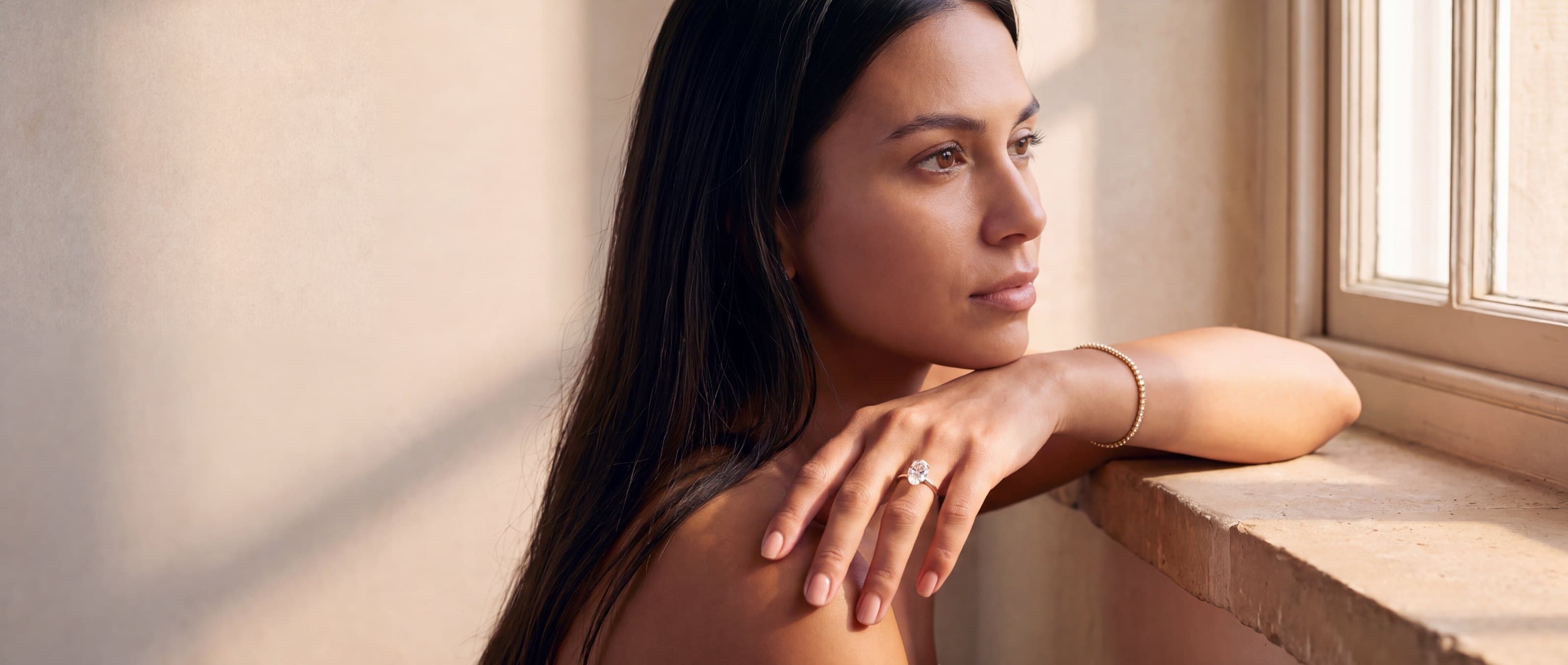 A woman by a sunlit window wearing an oval lab-grown diamond engagement ring and a diamond line bracelet