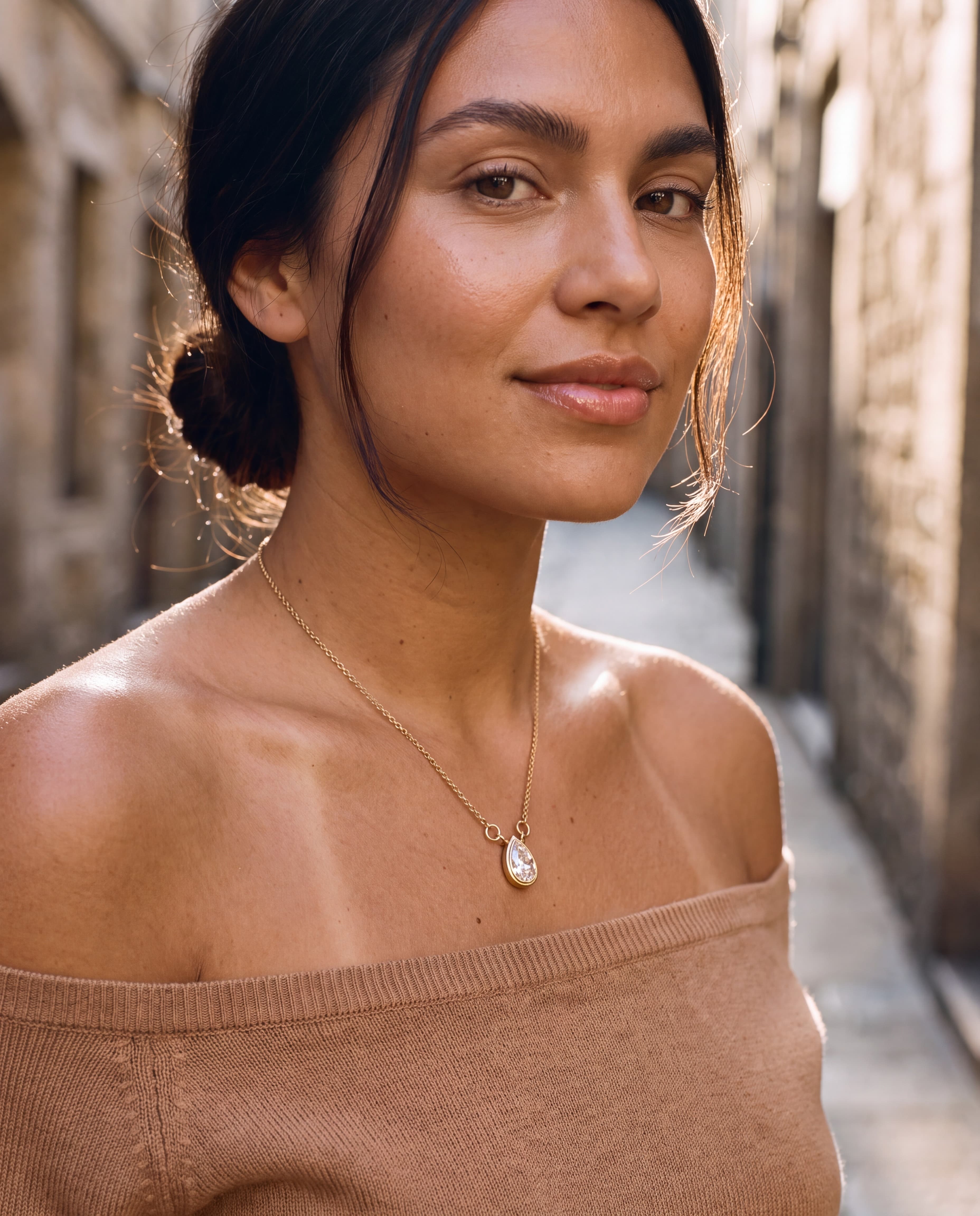 A sun-kissed young woman in an off-shoulder camel knit on a sunlit stone street, wearing a pear-shaped lab-grown diamond bezel pendant in 14K yellow gold