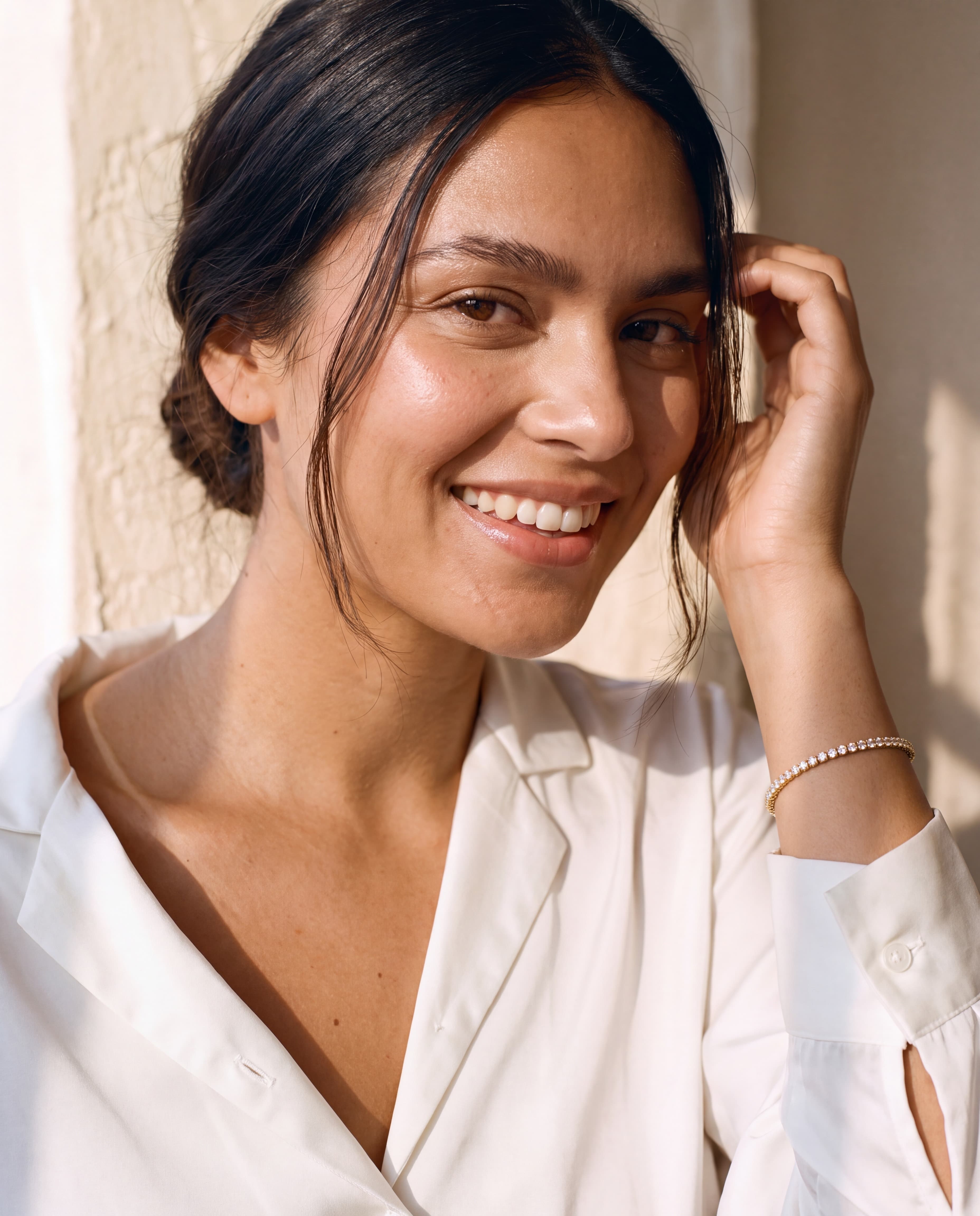 A sun-kissed young woman smiling as she tucks her hair behind her ear in an open-collared white silk shirt, wearing The Gabriella Line lab-grown diamond line bracelet in 14K yellow gold